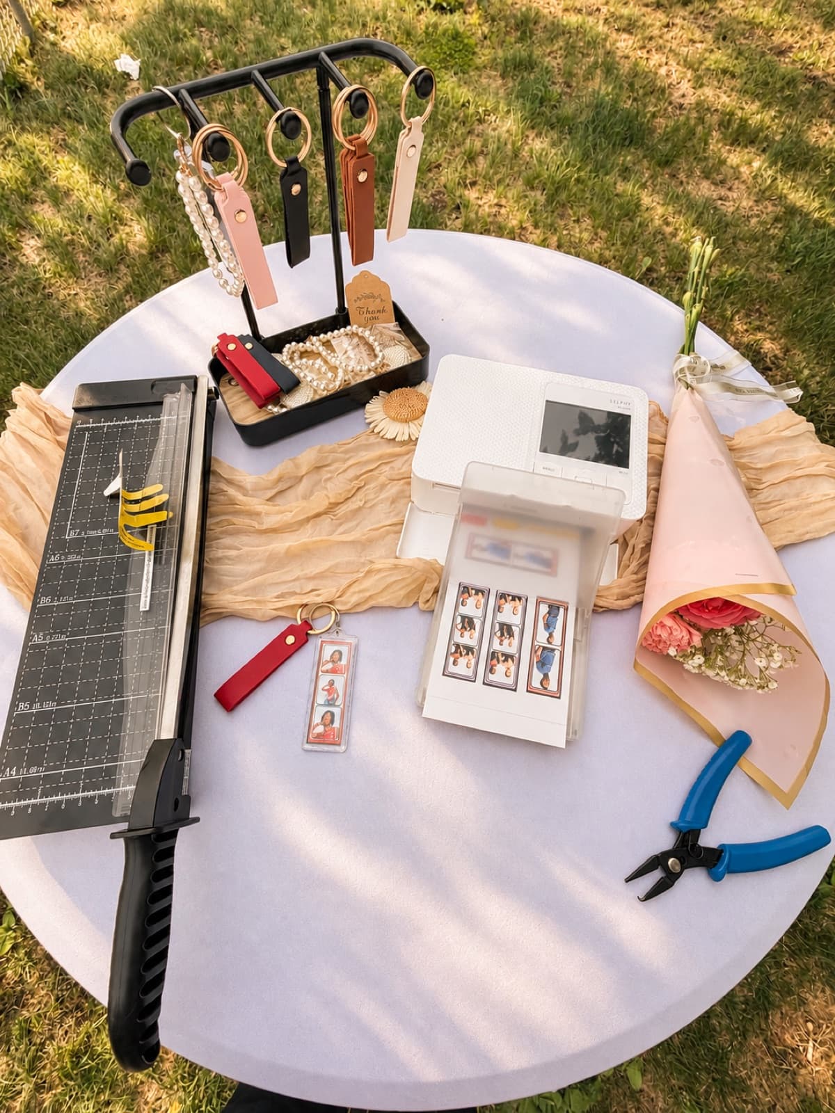 Photo keychain station on an outdoor table with a photo printer, trimmer, and keychains on a stand
