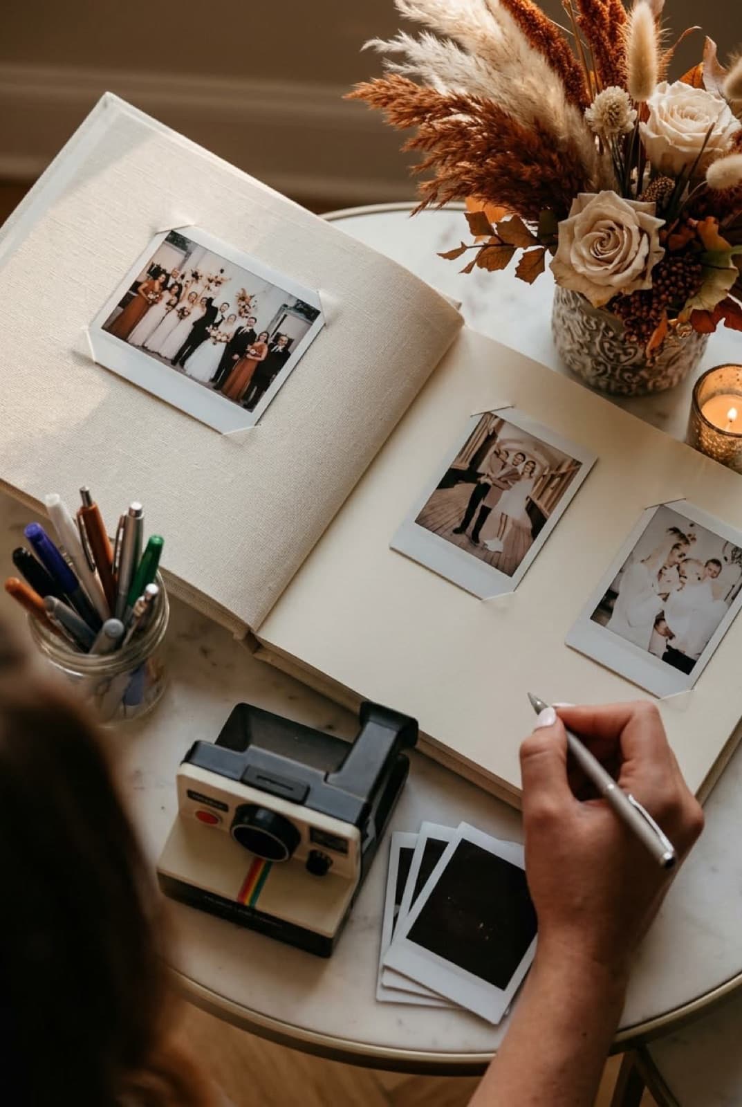 Guest writing a note beside instant photos in an open guest book, with a Polaroid camera and dried flowers