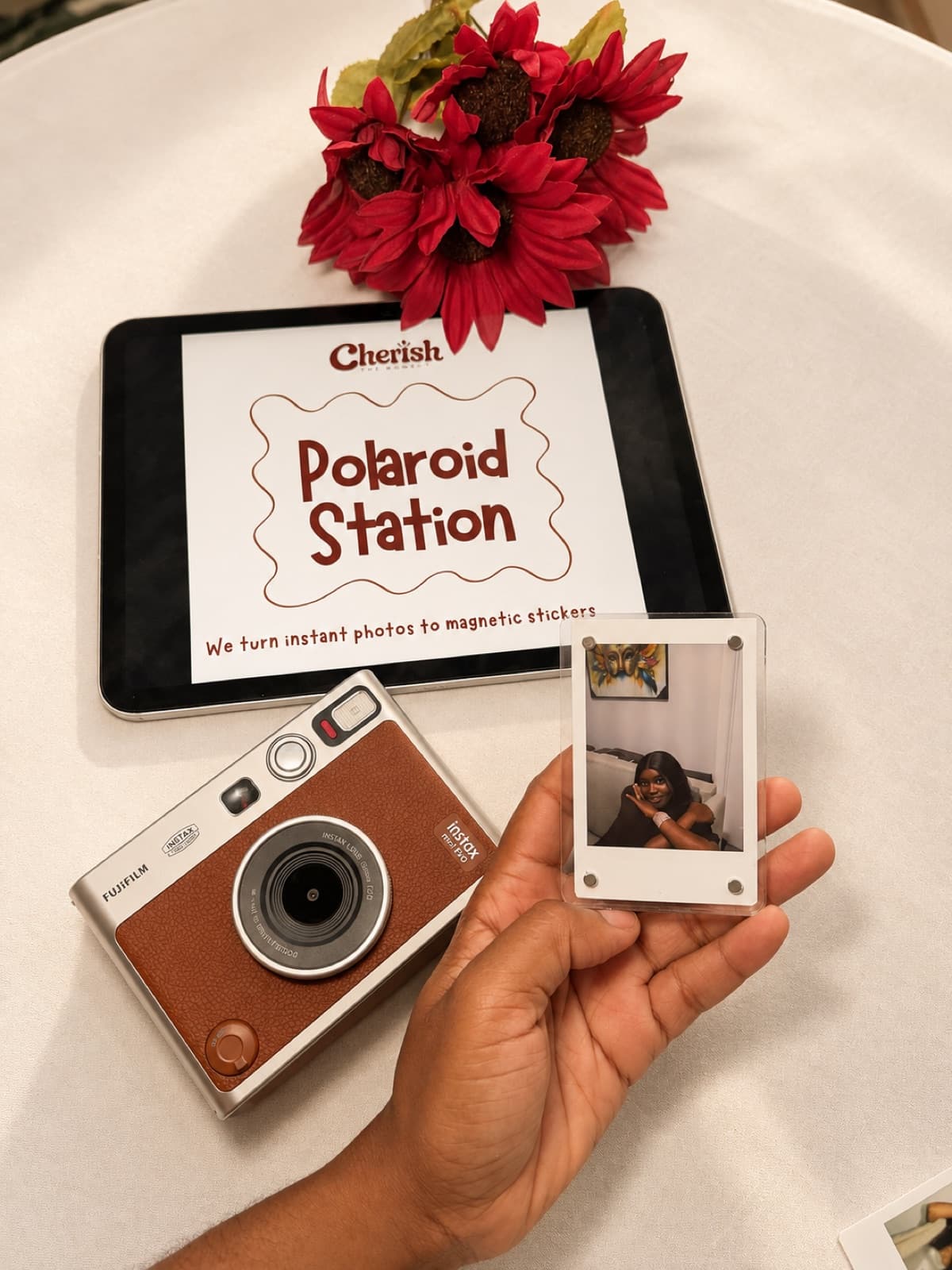 Hand holding a finished photo magnet in front of the Cherish Polaroid Station sign and instant camera